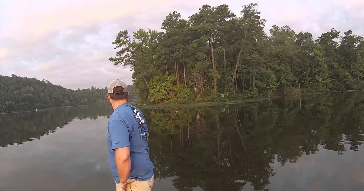 A man in a blue shirt and cap stands in a calm lake, surrounded by trees and a cloudy sky.