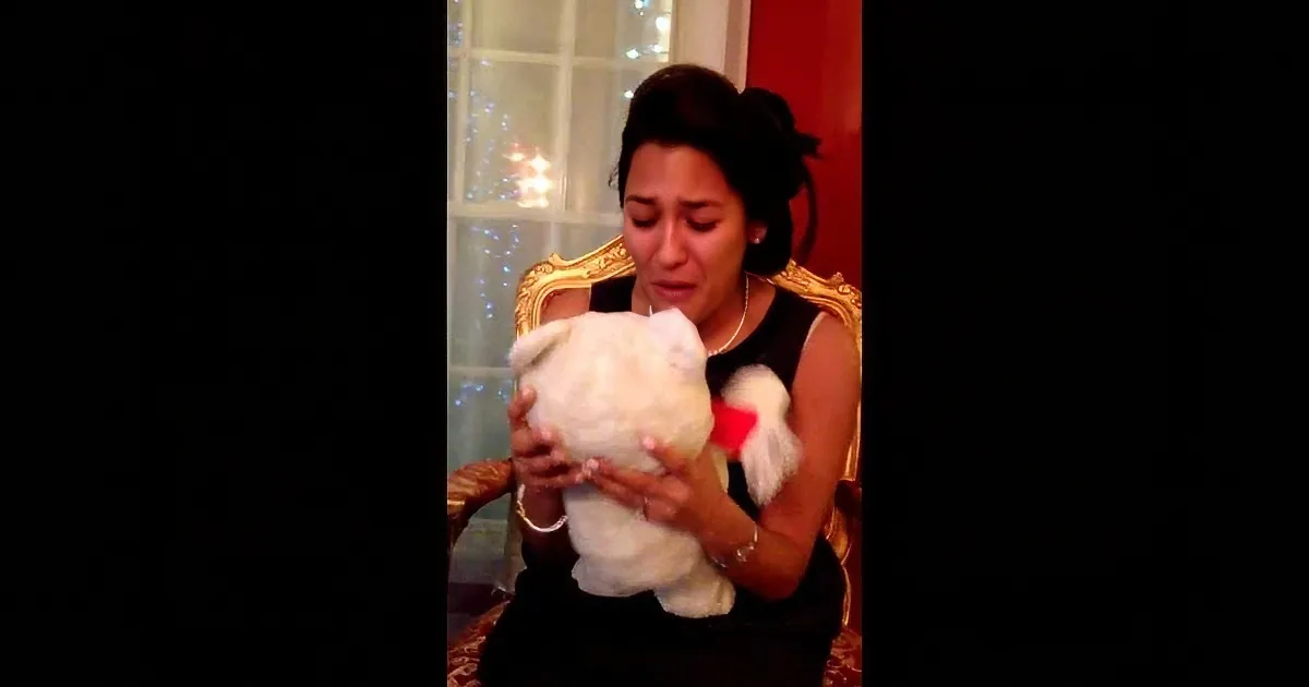 A woman in a black dress holds a white stuffed animal, appearing emotional, with a decorated window in the background.