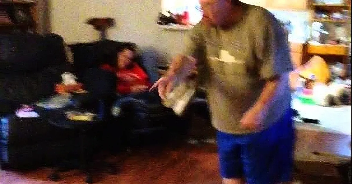 A man in a gray shirt and blue shorts stands in a living room, holding a white object, while a woman sits on a couch in...