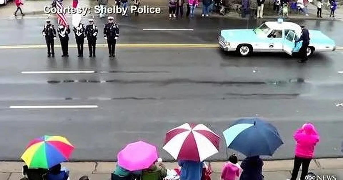 A police car drives past a line of uniformed officers and spectators holding umbrellas in the rain.