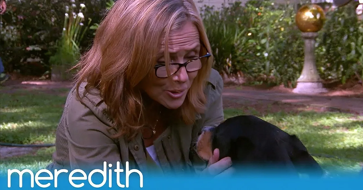 A woman with glasses holds a black dog in a garden setting, with "Meredith" text visible.