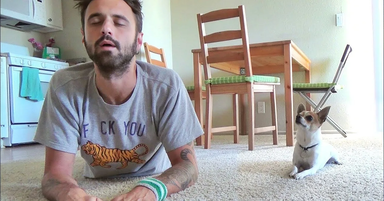 A man with closed eyes and a dog lying on the carpet in a cozy room with chairs and a kitchen.