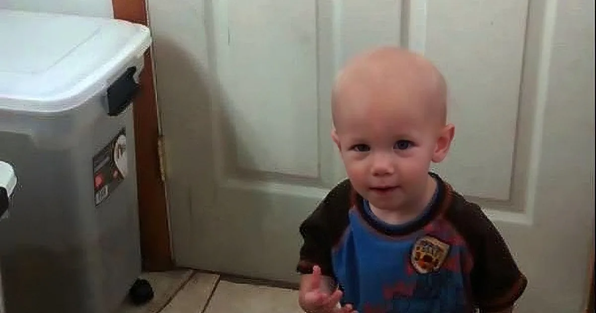 A young child with a bald head wearing a blue and brown shirt stands indoors near a door and a storage bin.