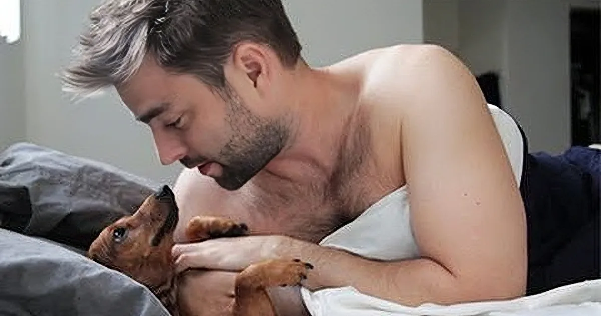 A shirtless man is lying in bed, holding and gently petting a small brown dog.