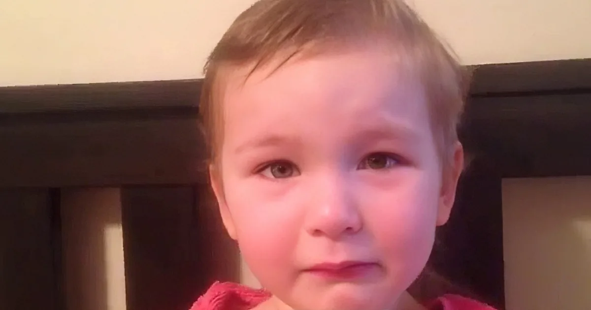 A young child with short hair, wearing a red top, sits in front of a dark wooden headboard, looking directly at the camera.