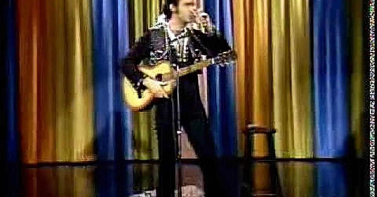 A man in a black suit plays a guitar and sings on stage with a blue curtain backdrop.