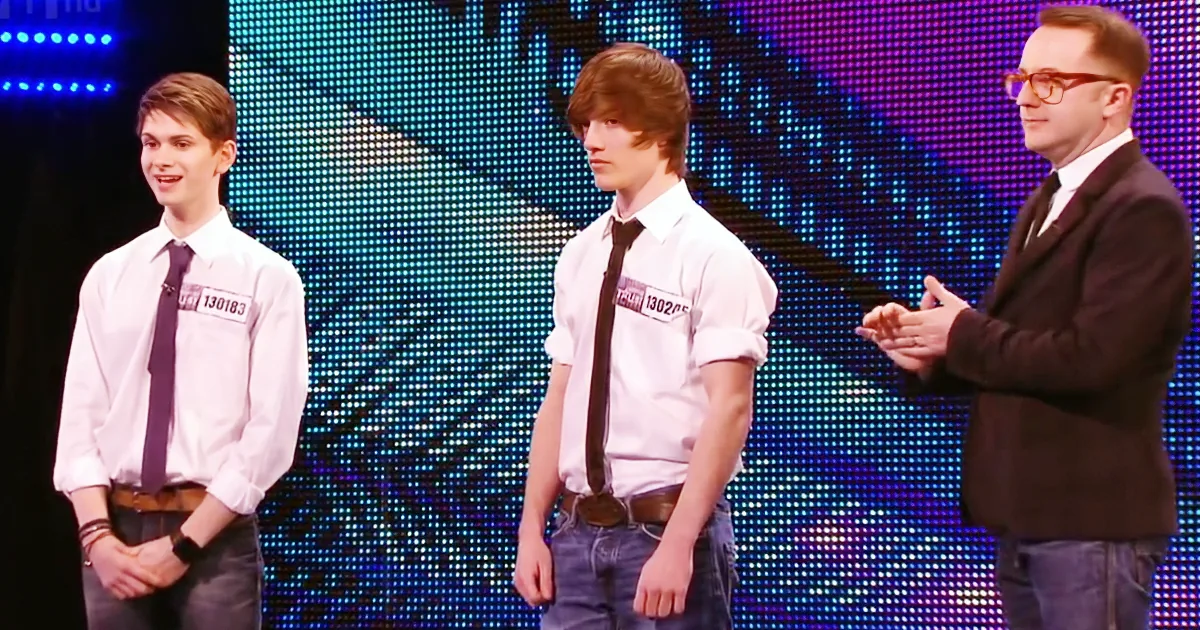 A man in a suit gestures to two young men in white shirts and ties on a stage with a colorful LED backdrop.