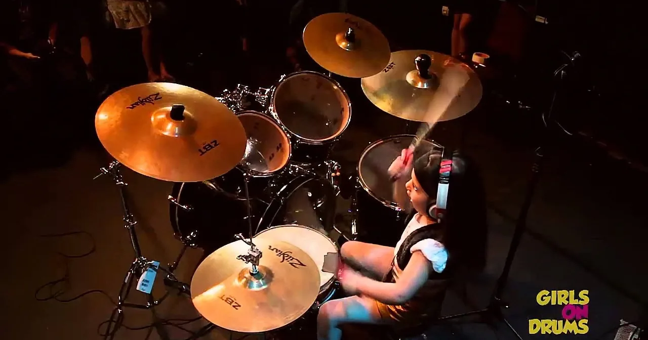 A young girl plays drums on stage with a cymbal set and drumsticks.