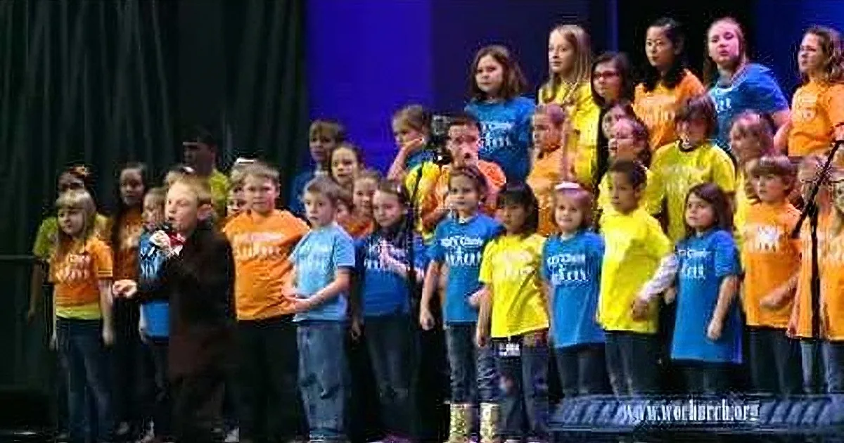 A man in a suit stands on stage with a group of children in colorful shirts, possibly singing or performing.