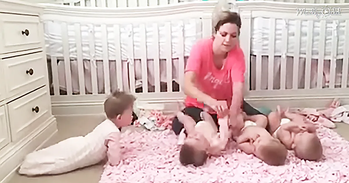 A woman in a pink shirt sits on the floor surrounded by four babies on a pink flower blanket.