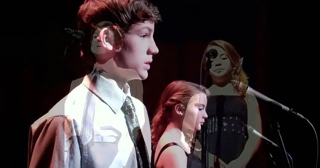 A young man in a brown robe sings into a microphone on stage, with other performers in the background.