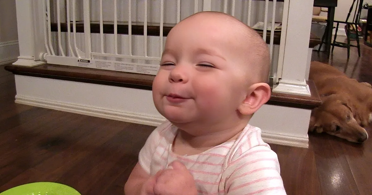 A baby in a striped shirt smiles while holding a green bowl, with a dog lying on the floor in the background.