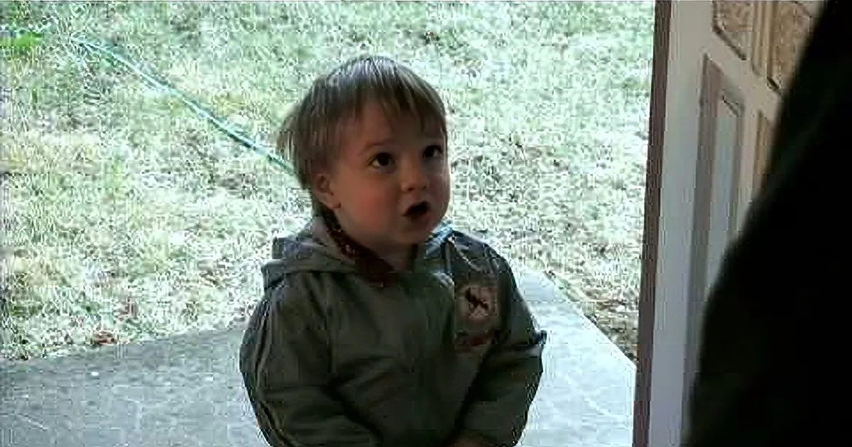 A young child in a green jacket stands outside, looking up with a surprised expression.