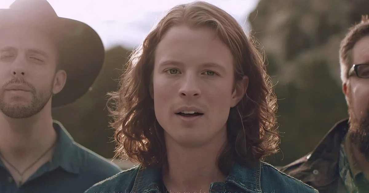 A person with long, wavy hair and a denim jacket stands in front of a group of men, one wearing a cowboy hat.
