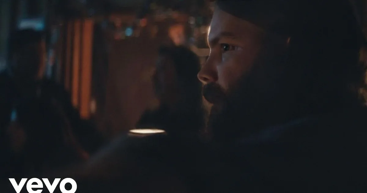 A man with a beard and dark hair is in a dimly lit room, looking to the side.
