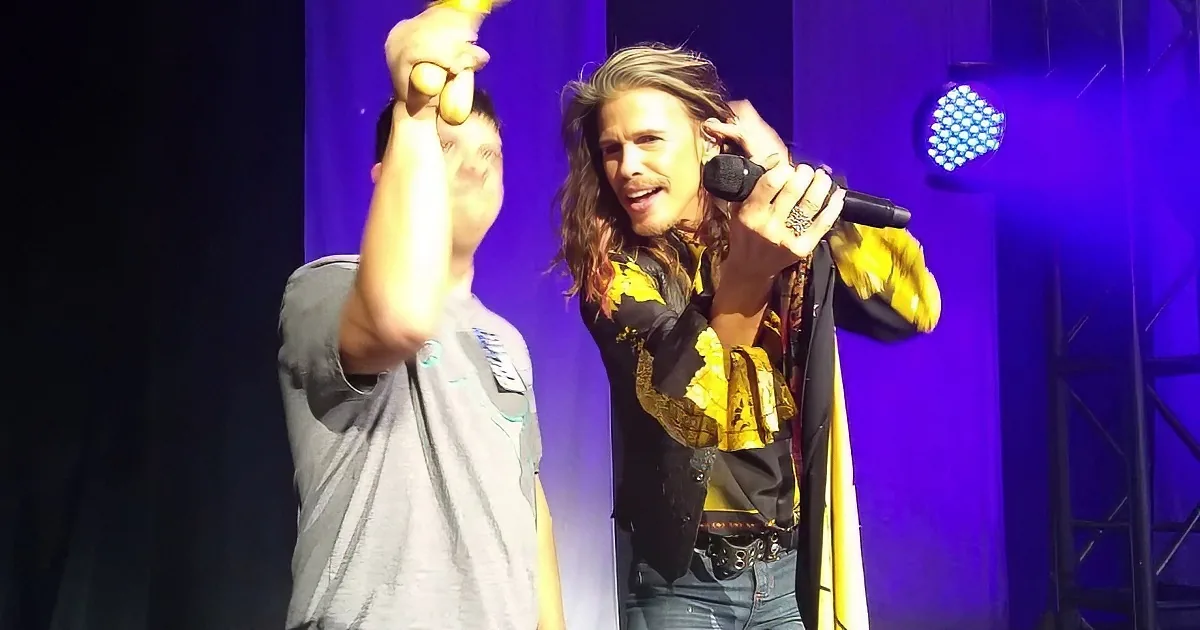 A man with long hair and a yellow jacket sings into a microphone on stage.