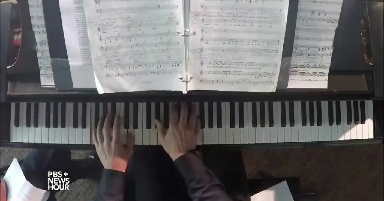 A person plays piano with sheet music open, hands positioned on keys.