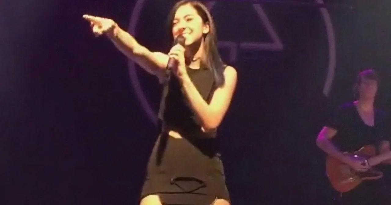 A woman in a black dress sings into a microphone, pointing to the left, with a guitarist in the background.