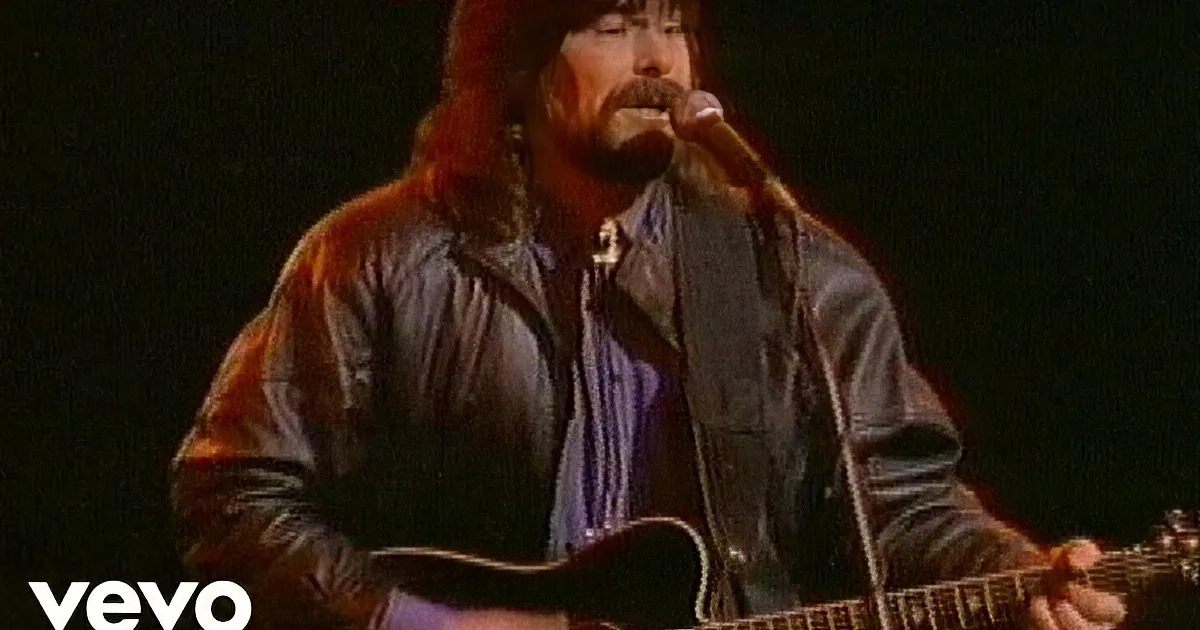A man with long hair and a beard sings into a microphone while playing a guitar.