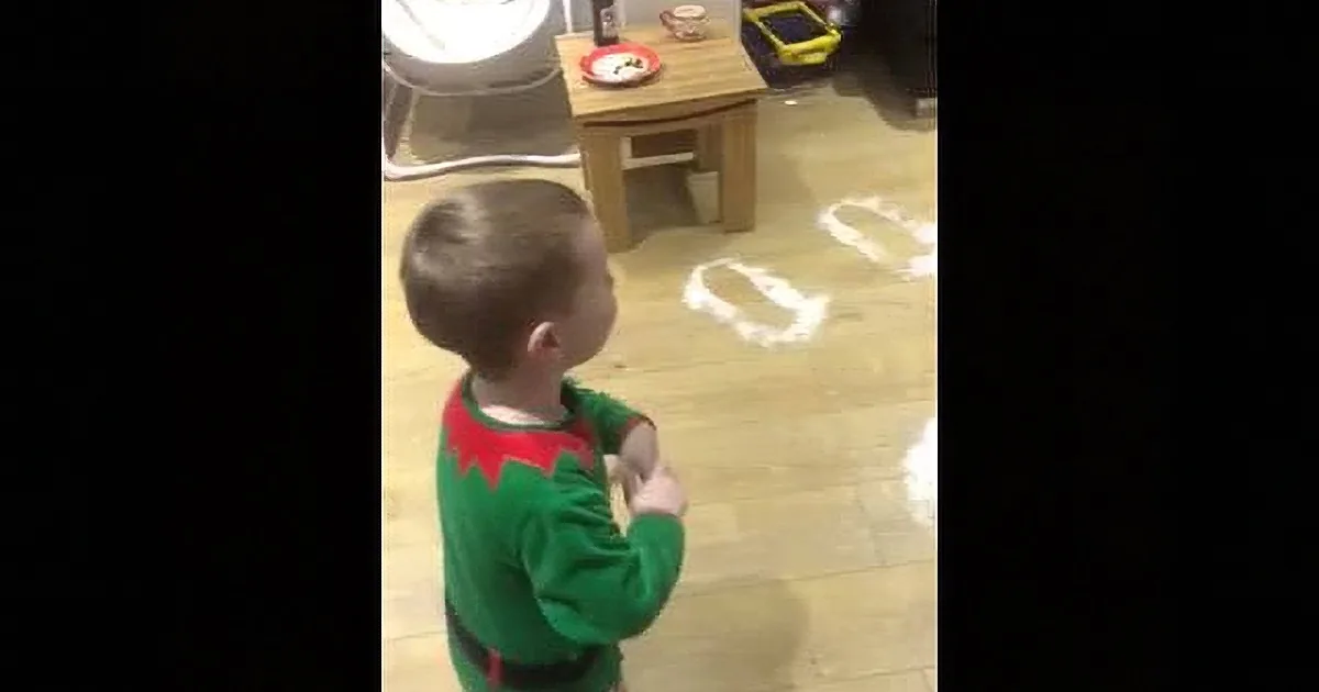 A child in a green Christmas outfit stands in a room with a small table and white footprints on the floor.