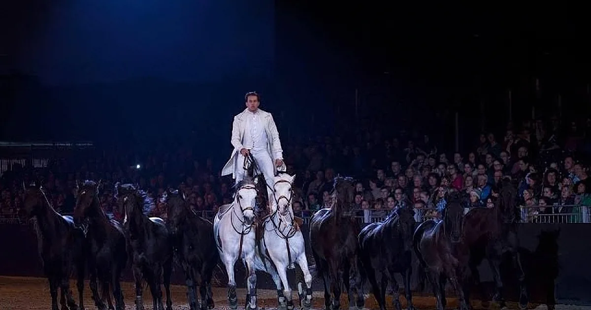 A man in white attire rides a white horse, leading a group of black horses in a dimly lit arena.