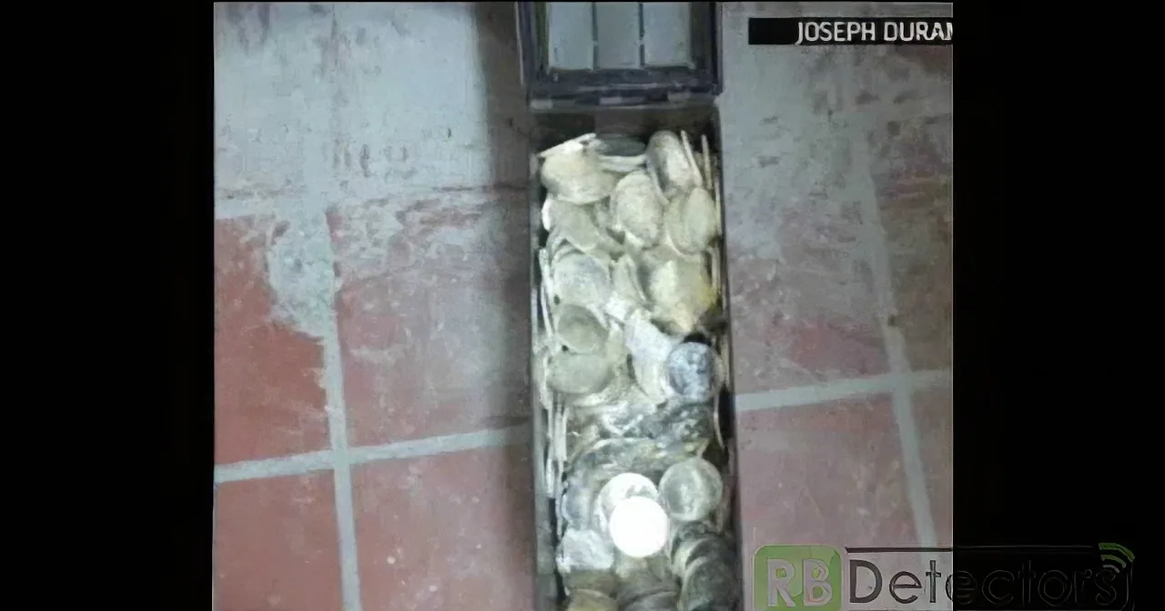A metal box filled with coins is placed on a brick surface.