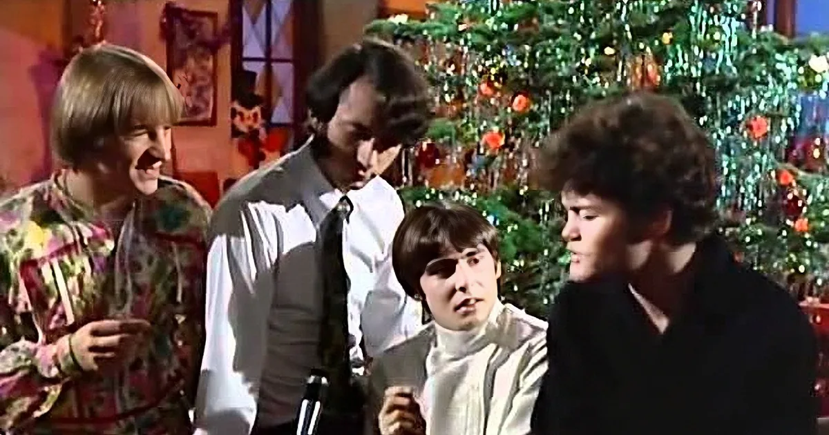 A man in a white shirt and tie stands beside a Christmas tree, surrounded by others in festive attire.