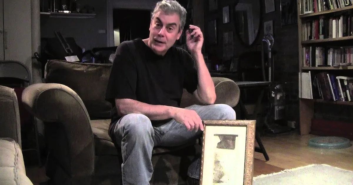 A man sits on a couch, holding a framed object, with a bookshelf and mirror in the background.