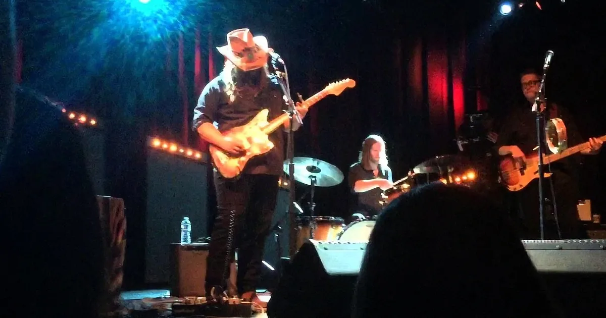 A guitarist in a cowboy hat plays a guitar on stage with a band behind him.