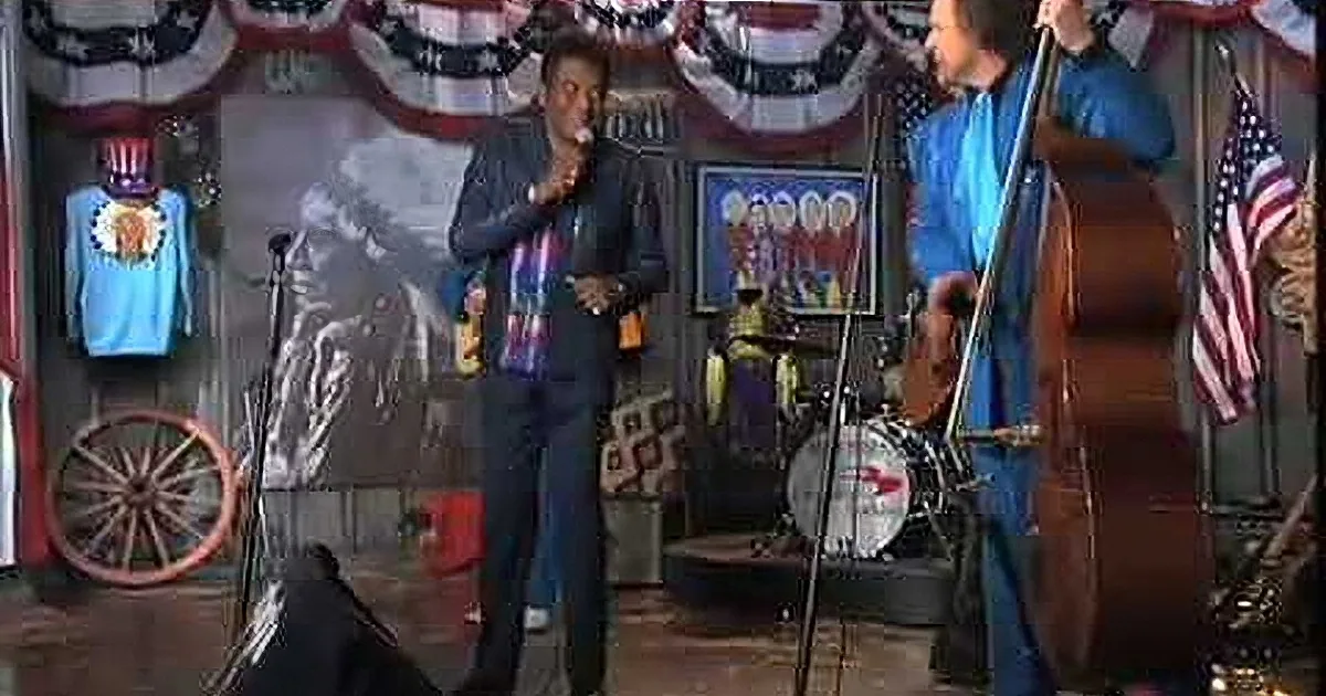 A man in a suit stands on stage, singing, with a band playing instruments behind him.