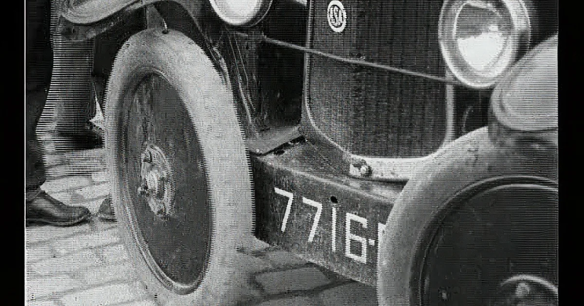 An old car with a visible license plate and a person standing nearby on a brick pavement.