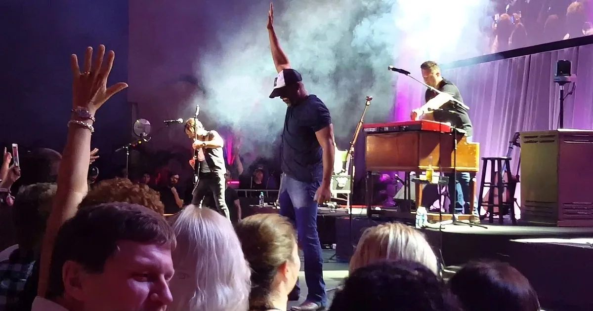 Musicians performing on stage with audience raising hands, smoke, and colorful lighting.