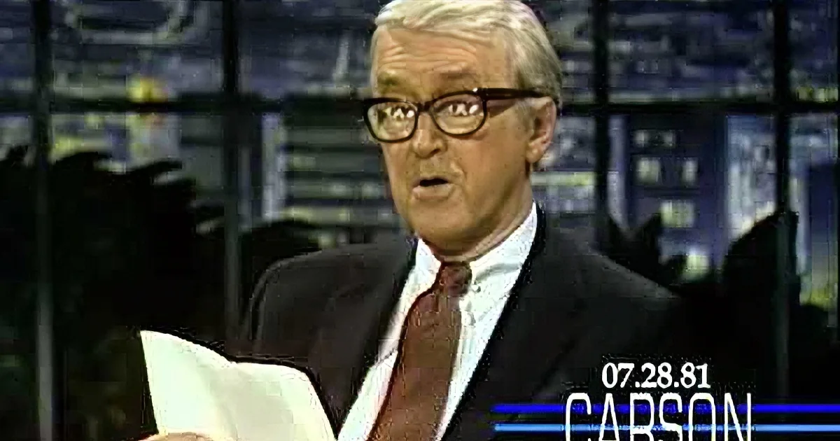A man in a suit and glasses holds a piece of paper, appearing to speak or read.