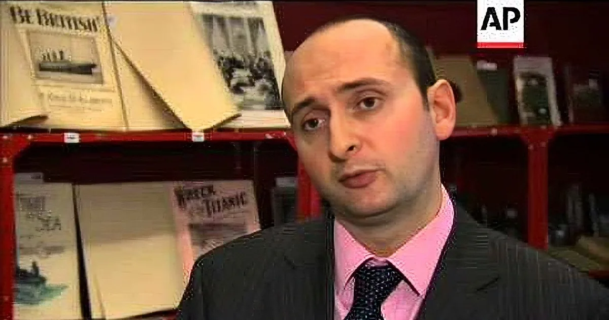 A man in a suit and tie is speaking, with a bookshelf of maritime-themed books behind him.