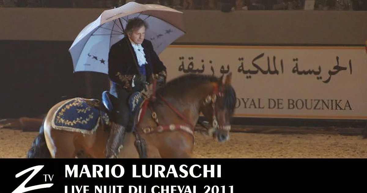 A man in formal attire rides a horse under an umbrella in a sand arena.