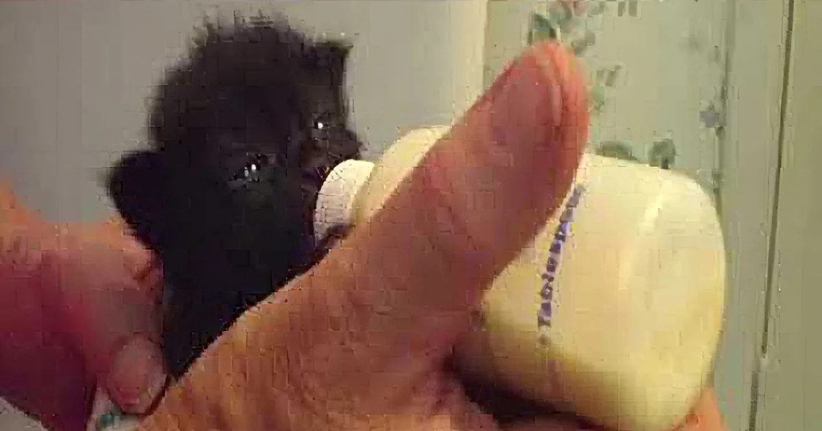 A tiny black kitten is being bottle-fed by a person's hand, with a bottle and a thumb visible.
