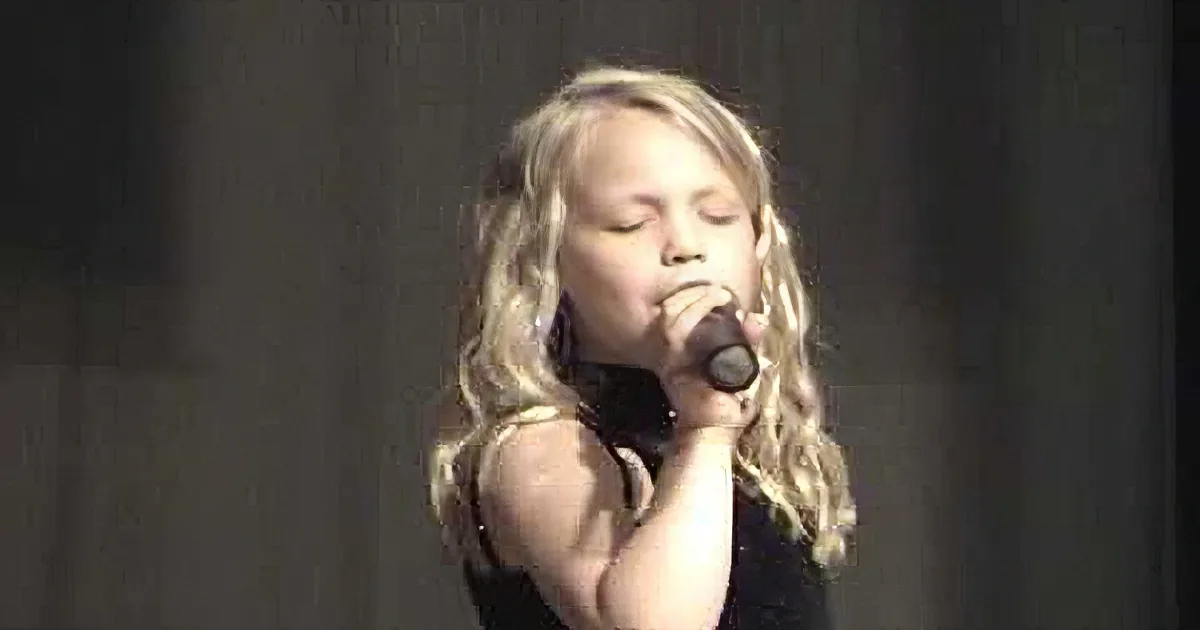A person with blonde hair singing into a microphone, wearing a black top, against a dark background.