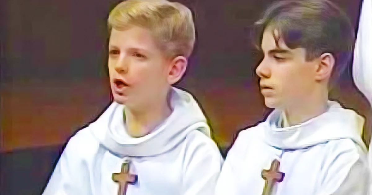 Two young boys in white robes with crosses, possibly singing or speaking, in a church setting.