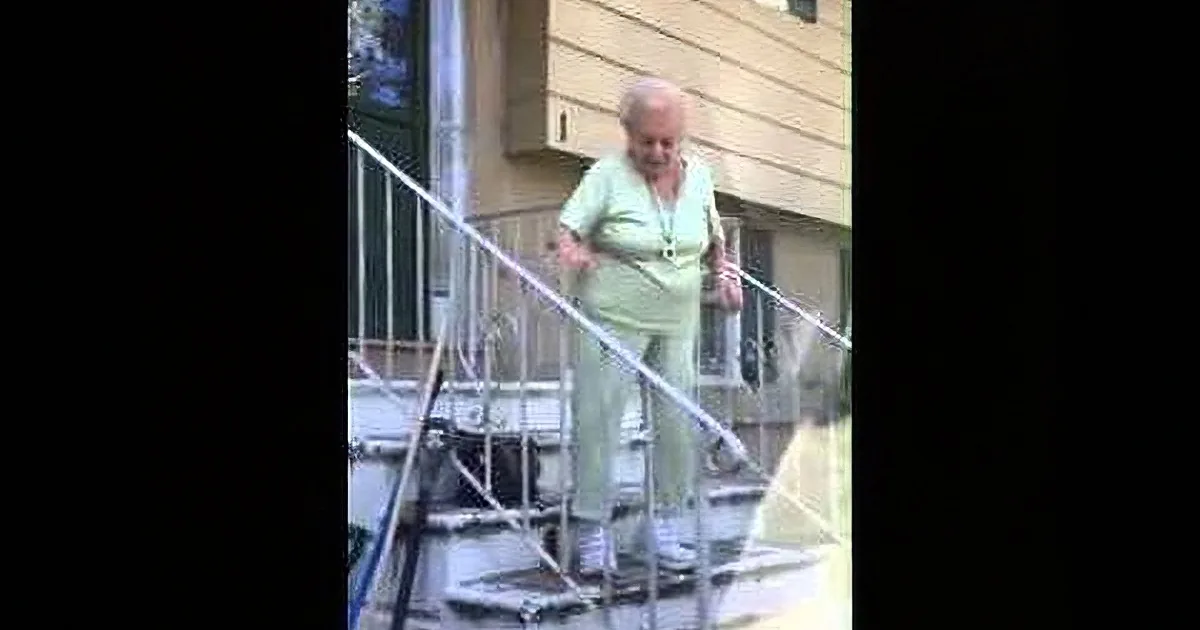 An elderly woman in a light green outfit is walking up a set of stairs, holding onto the railing.