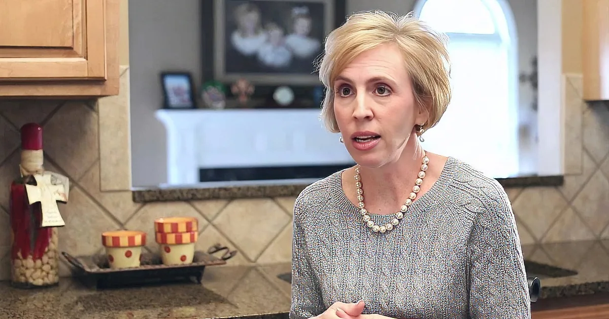 A woman in a gray sweater and pearl necklace stands in a kitchen, gesturing with her hands.