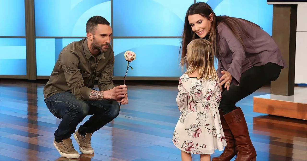 A man kneels, offering a rose to a young girl, while a woman watches nearby on a TV show set.