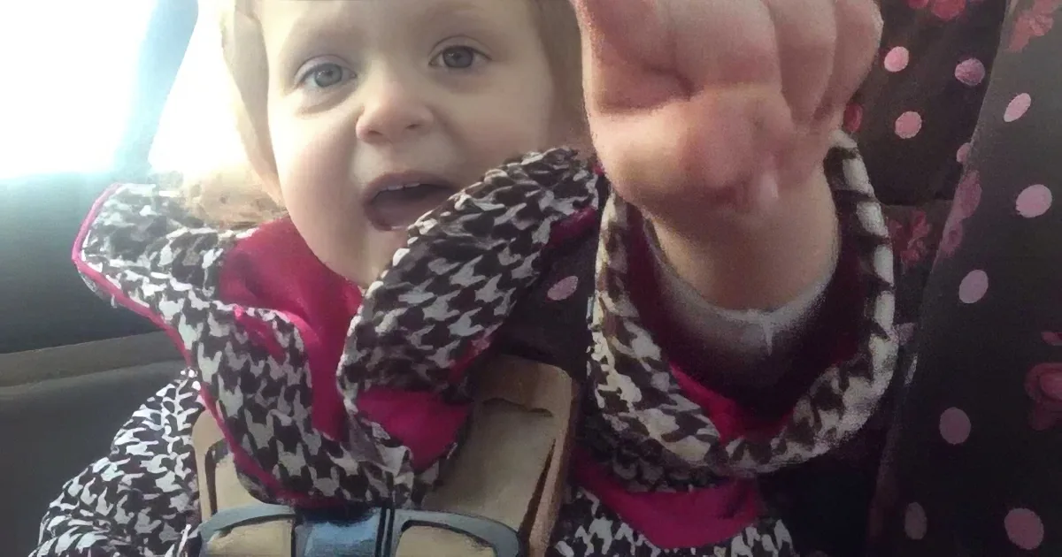 A baby in a car seat, wearing a patterned jacket, reaches out with one hand.