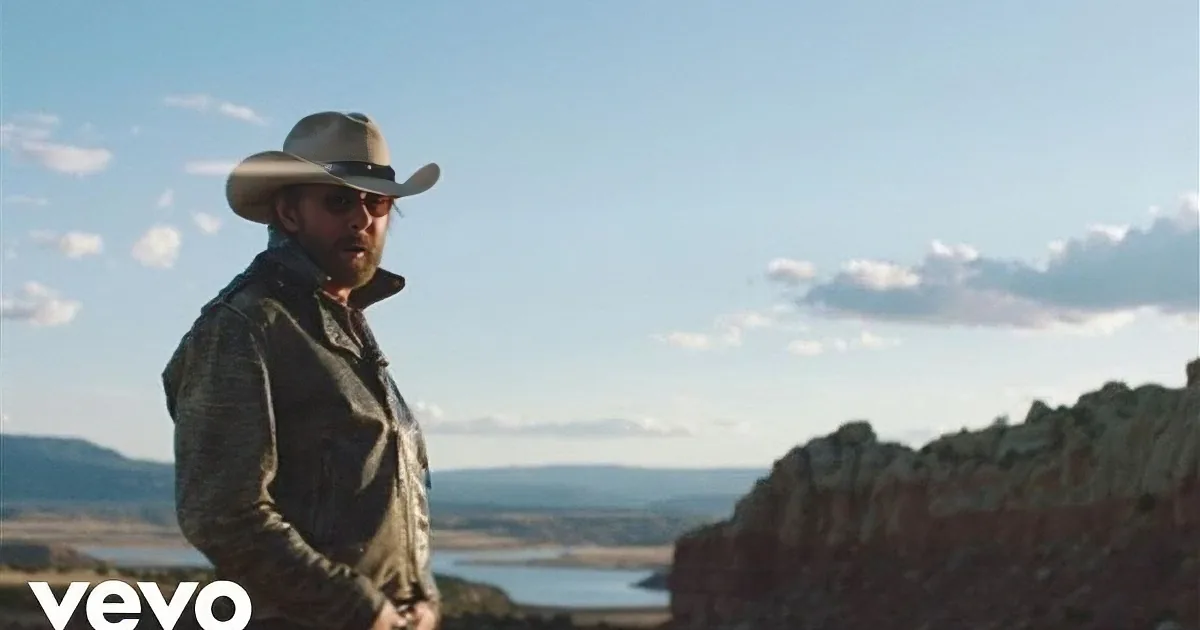 A man in a cowboy hat stands against a backdrop of a vast, rugged landscape with a body of water and a clear sky.