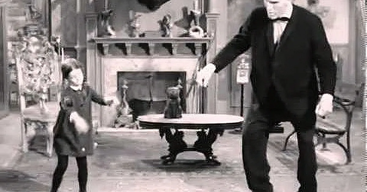 A man in a suit and bow tie extends his hand towards a young girl in a dress, both in a room with a fireplace and...