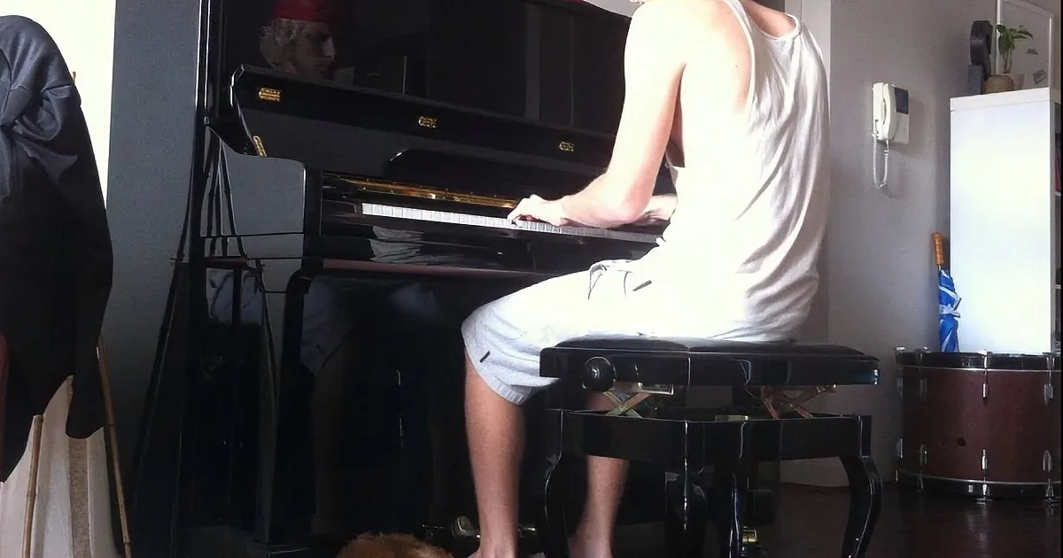 A person in a white tank top and shorts plays a black piano in a room with a drum set and a cat.
