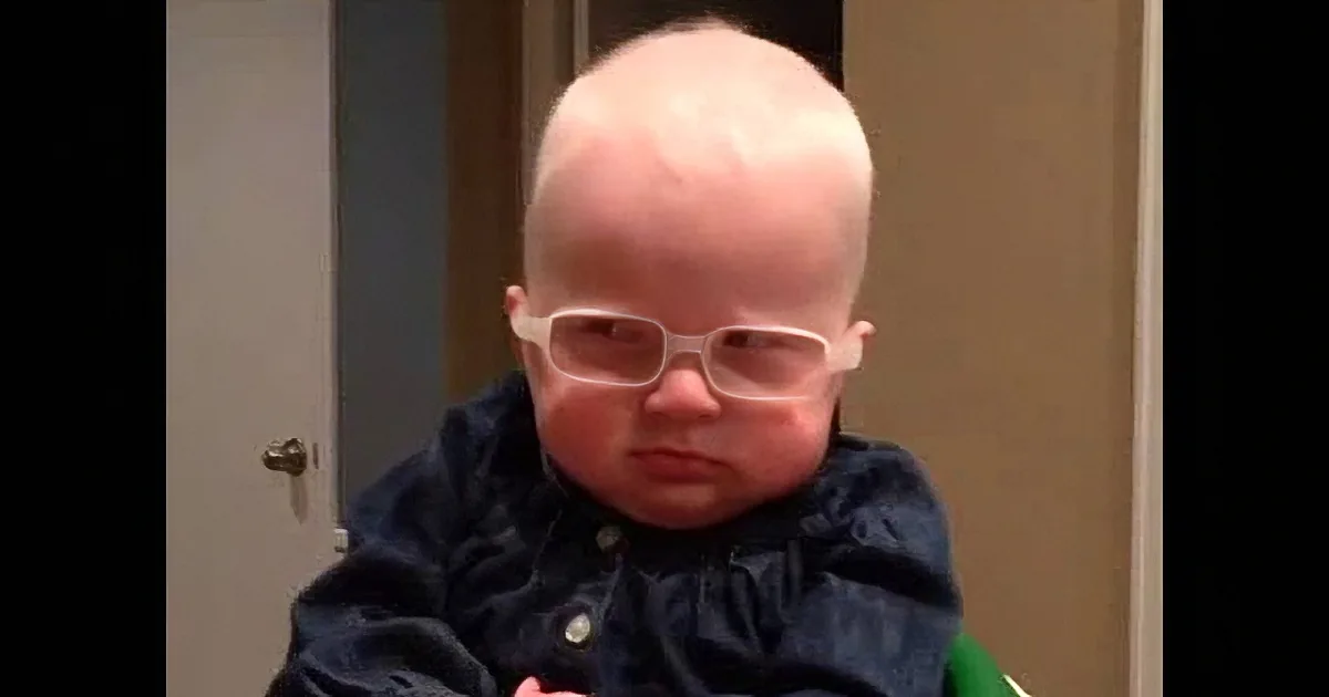 A baby wearing glasses and a dark jacket sits in a room with a door and wall visible.