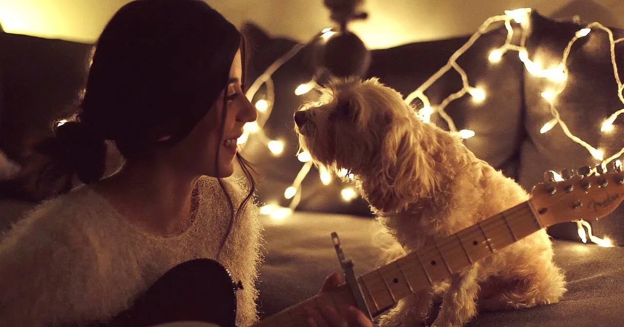 A woman smiles while playing guitar, her dog watches attentively under string lights.