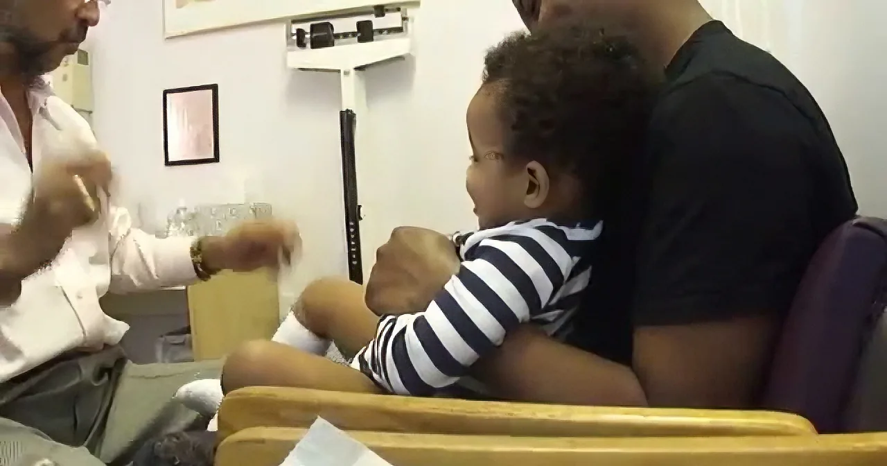 A baby in a striped onesie sits on a man's lap in a medical setting.