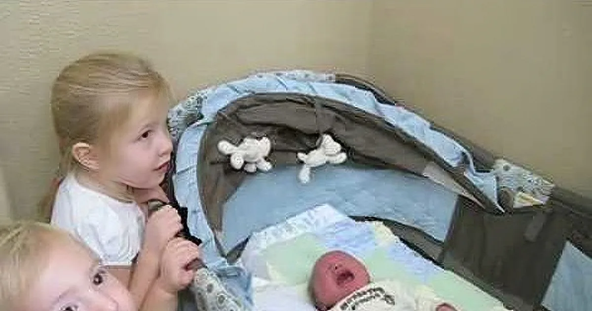 A young child gazes at a baby in a crib, surrounded by toys and a blanket.