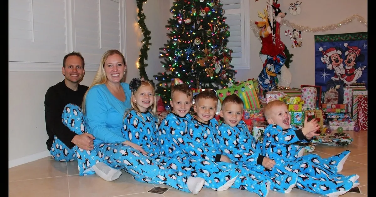 A family of seven in matching blue penguin pajamas poses in front of a Christmas tree and presents.